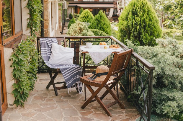 Ein einladender Holztisch, flankiert von zwei Holzstühlen, ist auf einem Balkon positioniert. Die Szenerie ist umgeben von zahlreichen Pflanzen. Terrassenlounge im Landhausstil 