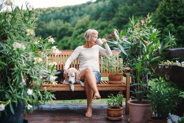 Ältere Dame sitzt auf gemütlicher Bank auf der Terrasse und trinkt Tee oder Kaffee